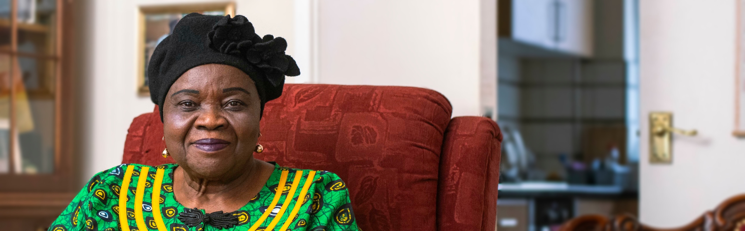 An older Black woman in traditional Nigerian dress sits in her living room, smiling at the camera