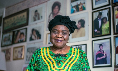 An older Black woman, with a wall of photographs behind her