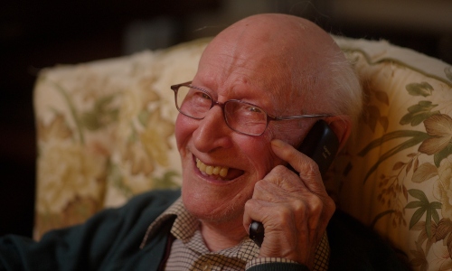 An older man with glasses chats on the phone, laughing
