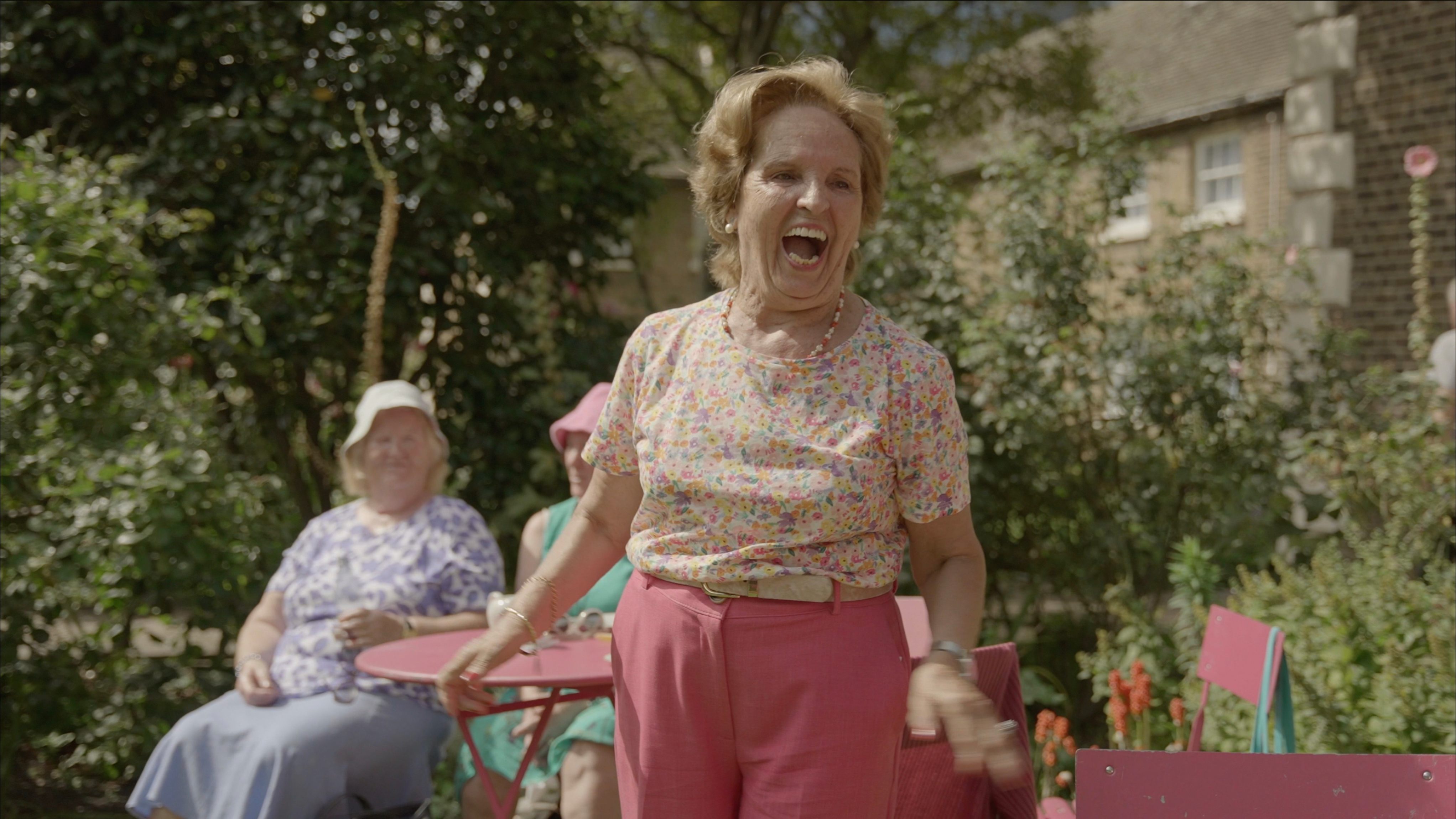 A woman in pink trousers and a large grin stands in a garden